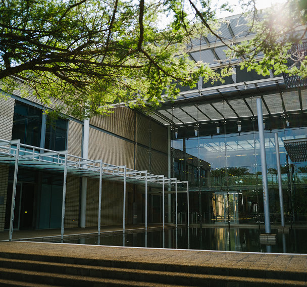Modern office building with glass entrance, reflecting pool, and overhanging trees.